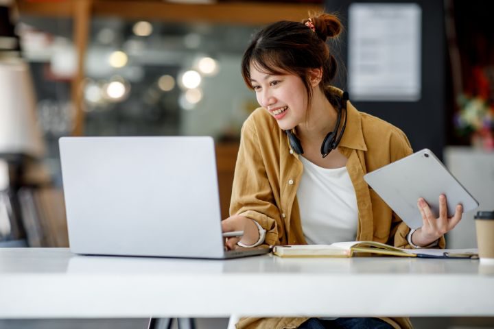 ヘッドフォンを首にかけた女性がノートパソコンを見ながら笑顔で勉強している画像