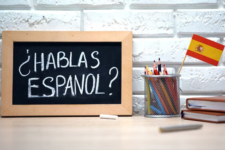机の上に2冊の本とスペイン語話せますかと書かれた黒板、スペインの旗が付いた棒の入ったペン立てが写っている画像