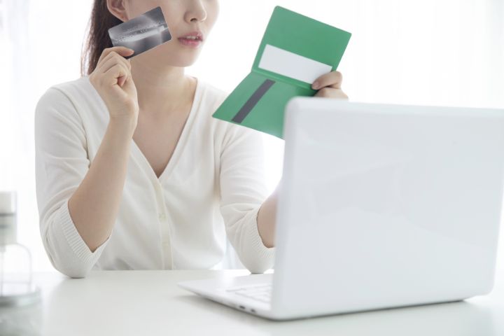 パソコンの前で通帳とカードを持っている人のイメージ
