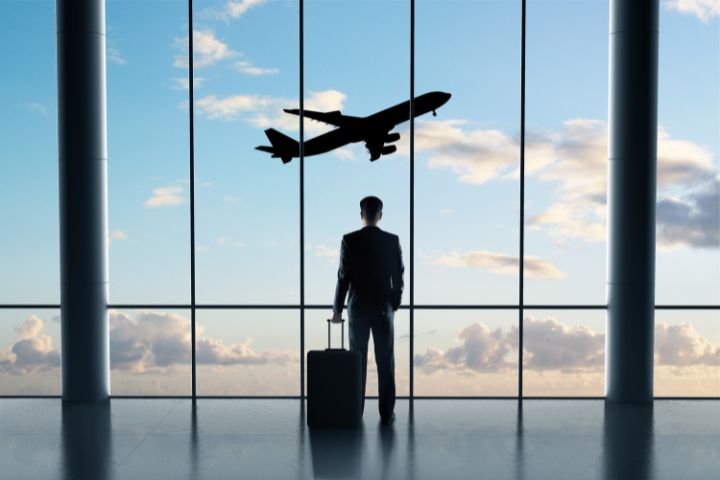 空港で飛んでいる飛行機を見ているスーツケースを持ったビジネスマンの画像